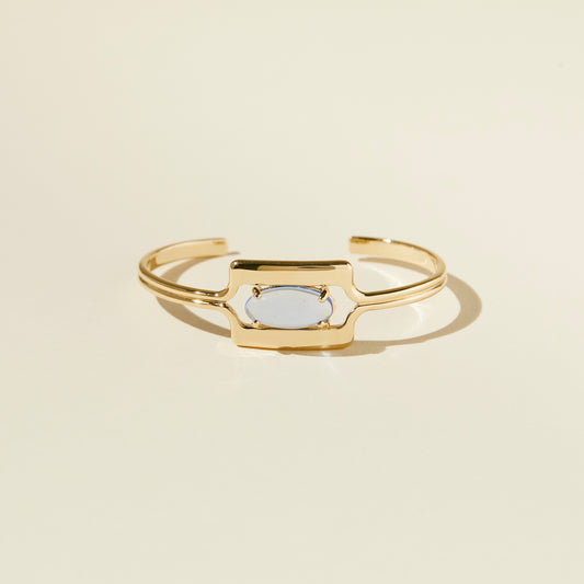 Lindsay Lewis Walton Cuff. Refined, smooth angles frame the elongated, prong set oval glass. Gold plated brass featuring a transluscent, sky blue, Czech glass adornment.